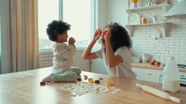 Küçük kızı kıvırcık saçlı Afro-Amerikalı kadın mutfakta hamur işi pişiriyor ve eğleniyor. Anne ve kızının kurabiye kesicilerle oynaması komik..