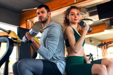 Spor kıyafetleri içinde ince bir kadın spor salonunda spor malzemelerinin arka planında dambıllarla antrenörle egzersiz yapıyor. Aktif sağlıklı yaşam tarzı, spor.