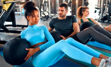Slim women african american and caucasian ethnicity and muscular man in sportswear doing exercises with a heavy medicine ball on a rubber mat in a gym club. The concept of sports and recreation.