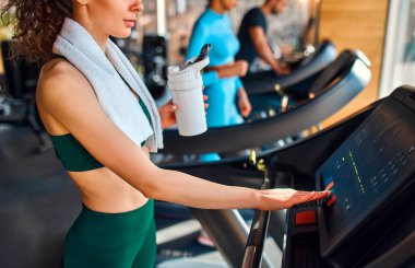 Spor salonunda koşu bandında koşan etnik çeşitliliğe sahip bir grup genç. Havlu taşıyan kızıl saçlı kız su içiyor ya da protein içeceği içiyor. Kardiyo çalışması. Spor ve eğlence kavramı..