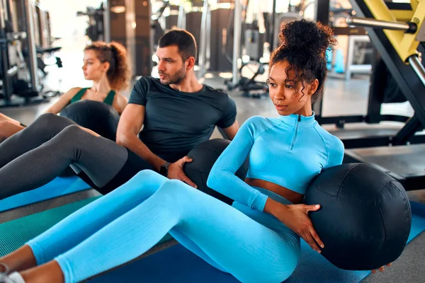 Zayıf Afrikalı kadınlar, beyaz ırk ve spor giyimli kaslı erkekler spor salonunda spor salonunda lastik bir paspasın üzerinde ağır bir ilaç topuyla egzersiz yapıyorlar. Spor ve eğlence kavramı..