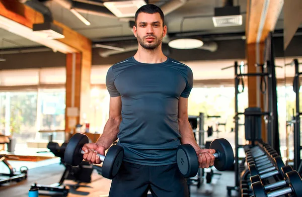 Spor giyimli kaslı atletik bir adam spor salonunda pazılar için halterlerle egzersiz yapıyor. Spor ve eğlence kavramı..