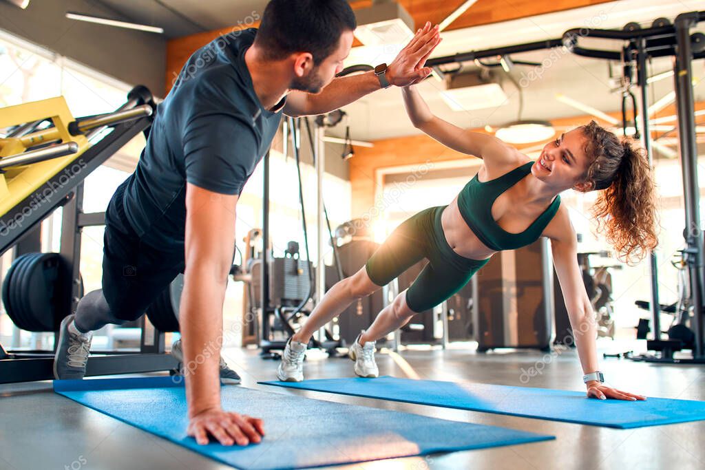 Slim woman and muscular man in sportswear doing plank exercise and high five on rubber mat in gym club. Athletic young couple. The concept of sports and recreation.