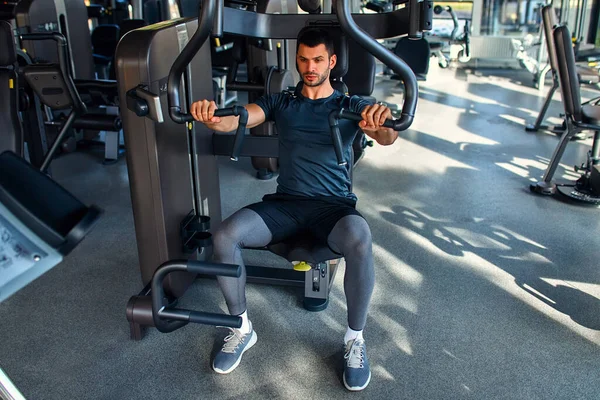 Spor giyim bölümündeki kaslı genç adam spor salonunda simülatör üzerinde egzersiz yapıyor. Spor ve eğlence kavramı..