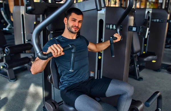 Spor giyim bölümündeki kaslı genç adam spor salonunda simülatör üzerinde egzersiz yapıyor. Spor ve eğlence kavramı..
