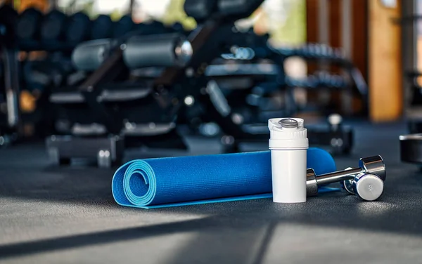 A rolled up blue yoga mat and a white metal flask for a bottle of water or a protein shake, next to it are dumbbells on the floor against the background of sports equipment in the gym.