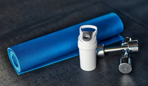 A rolled up blue yoga mat and a white metal flask for a bottle of water or a protein shake, next to it are dumbbells on the floor against the background of sports equipment in the gym.