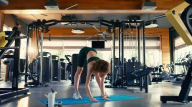 A slender athletic woman in sportswear does stretching exercises on a rubber mat in the club's gym against the background of sports equipment. Active healthy lifestyle, sports.