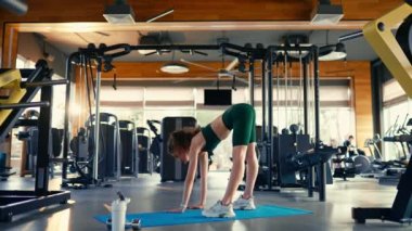 A slender athletic woman in sportswear does stretching exercises on a rubber mat in the club's gym against the background of sports equipment. Active healthy lifestyle, sports.