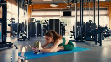 Spor kıyafetleri içinde ince bir atletik kadın spor malzemelerinin arka planına karşı kulübün spor salonundaki lastik bir paspasın üzerinde otururken esneme egzersizleri yapıyor. Aktif sağlıklı yaşam tarzı, spor.