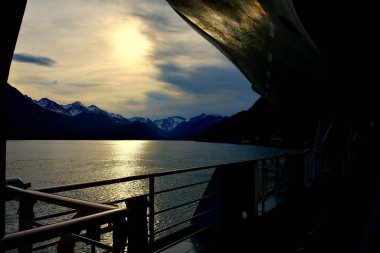Akşamları deniz koyunda dağlar ve ön planda gemi rayları.