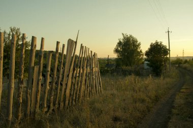 Kırsal bir yol boyunca uzanan harap olmuş ahşap bir çit. Bir yaz akşamı gün batımında.