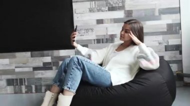 Açık renk ceketli güzel bir kız evde bir sandalyede oturuyor ve cep telefonuyla video bağlantısı ile konuşuyor..