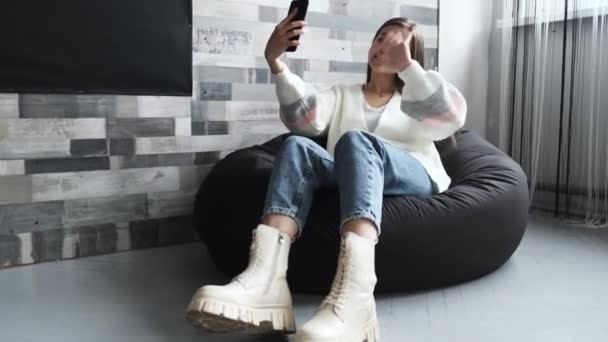 Egy csinos fiatal lány fénykabátban ül otthon egy széken, és videokapcsolaton keresztül beszél egy mobiltelefonon keresztül.. — Stock videók