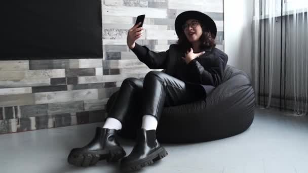 Egy csinos fiatal lány fekete kalapban ül otthon egy széken, és videokapcsolaton keresztül beszél egy mobiltelefonon keresztül.. — Stock videók