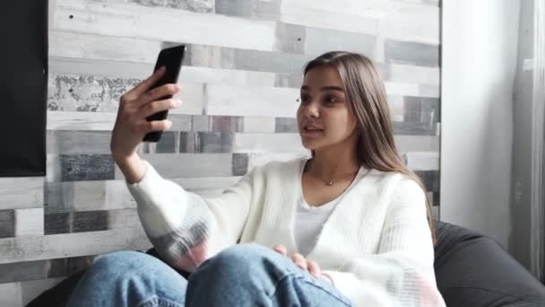 Una chica muy joven en una chaqueta ligera está sentada en una silla en casa y hablando a través de un enlace de vídeo a través de un teléfono móvil. — Vídeos de Stock