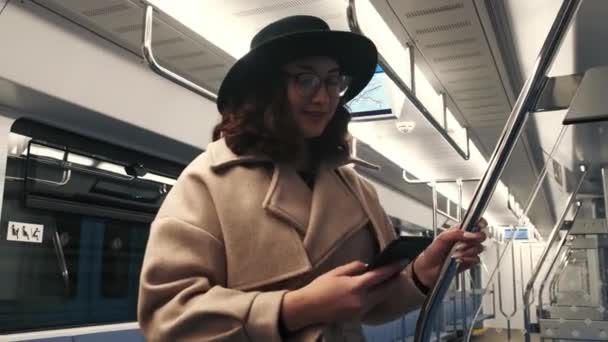 Una joven bonita con un abrigo y un sombrero negro está sentada en el teléfono mientras está en un vagón del metro. Un vagón de metro vacío sin gente. — Vídeo de stock