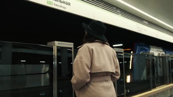 Achteraanzicht van een jong meisje met een hoed naast een passerende metro wachtend op een lift. — Stockvideo