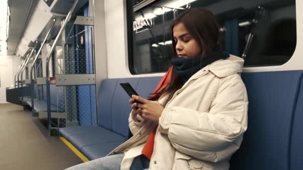 En söt ung flicka i en färgad halsduk sitter i telefonen i en tunnelbanevagn. En tom tunnelbanebil utan människor. — Stockvideo