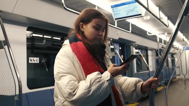 Seorang gadis muda yang cantik dengan syal berwarna sedang duduk di telepon saat di kereta bawah tanah. Sebuah kereta bawah tanah yang kosong tanpa orang. — Stok Video