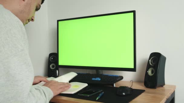 Por cima do ombro está uma imagem de um tipo a trabalhar em casa num computador numa secretária, a olhar para uma tela verde e a fazer anotações no seu caderno. — Vídeo de Stock