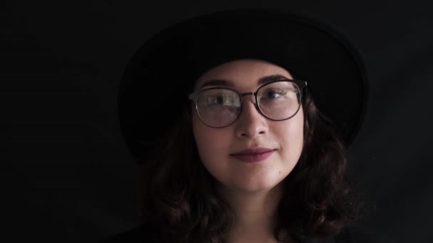 Retrato de cerca de una chica bonita con un sombrero sobre un fondo negro, una mujer joven y elegante mirando a la cámara. Los arribistas. Moda, belleza. Retratos femeninos. — Vídeo de stock