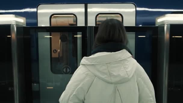Widok z tyłu młodej dziewczyny w białej kurtce czekającej na peronie stacji metra, wchodzi do pustego powozu. — Wideo stockowe