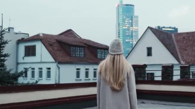 Uzun beyaz saçlı ve beyaz şapkalı bir kız şehrin manzarasına bakıyor. Yavaş çekim. Kızlardan şehrin manzarası.