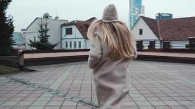 Paltolu ve beyaz şapkalı uzun saçlı bir kız şehirde ağır çekimde koşuyor. Arka planda şehir ve iş merkezi var. Yavaş çekim