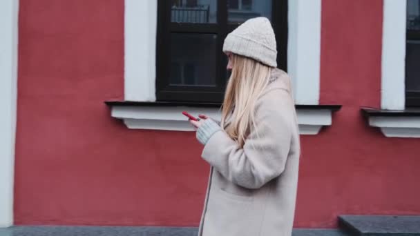 白い帽子と美しいコートでファッショナブルな学生、イベントやソーシャルメディアに追いつくために大学に行く途中で彼女のスマートフォンで彼女のニュースフィード。The — ストック動画
