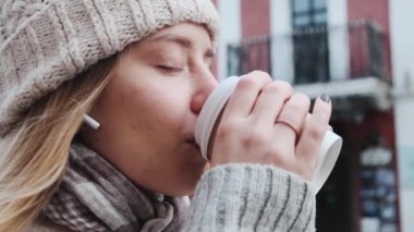 Beyaz şapkalı ve güzel ceketli genç bir kız, kablosuz kulaklık kullanarak müzik dinliyor ve arka planda ağır çekimde kahve içiyor.