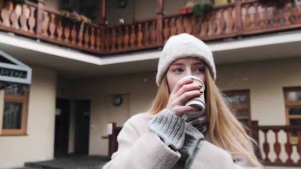 白い帽子と美しいコートの若い女の子は、ワイヤレスヘッドフォンを使用して音楽を聴き、ゆっくりと動きながらコーヒーを飲みます。 — ストック動画