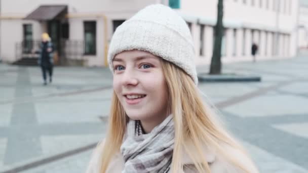 Close-up van een jong lachend meisje met wit haar met een hoed en sjaal. Portret van een meisje tegen de drukte van de stad. — Stockvideo