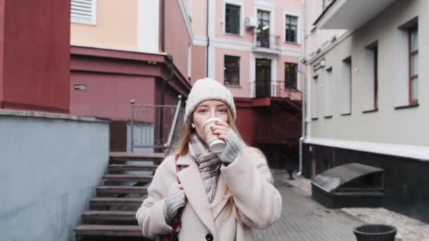 A young beautiful blonde in a white coat walking through town, listening to music and drinking coffee. Slow motion footage of a stroll through the city. — Stock Video