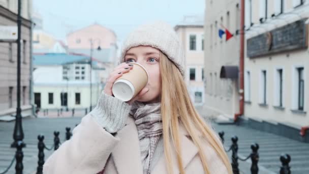 Primer plano de una joven rubia bebiendo café caliente contra el telón de fondo de la ciudad en cámara lenta. Una chica esperando amigos, colegas, familia. — Vídeo de stock