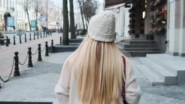 Επιστροφή άποψη ενός κοριτσιού με ξανθά μαλλιά, περπατώντας κάτω από ένα δρόμο στο κέντρο της πόλης κοιτάζοντας γύρω από τα φώτα της πόλης στο παρασκήνιο. Κοντινή αργή κίνηση — Αρχείο Βίντεο