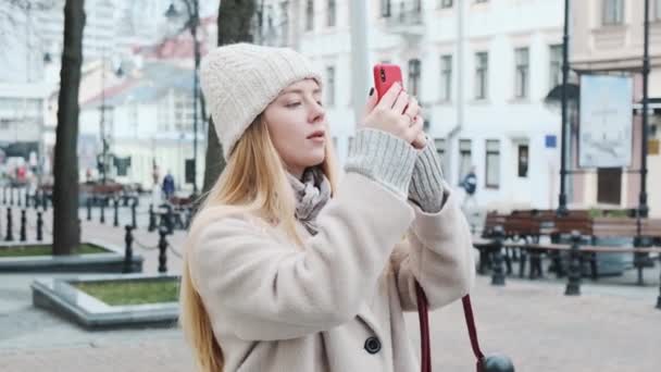 A fashionable student in a white hat and a beautiful coat, shooting a video on her smartphone for social media. Walking through a beautiful city. — Stock Video