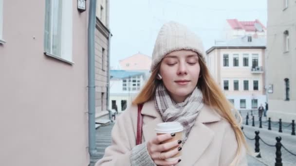 Primer plano de una joven rubia paseando por la ciudad, escuchando música y tomando café en cámara lenta, durante la temporada navideña. — Vídeo de stock