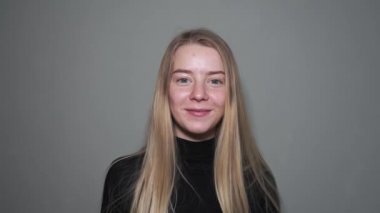 Gri arka planda, parmaklarını kaldıran ve gülümseyen sarışın bir kızın yakın çekimi. Parlak ve ışıltılı duygular.