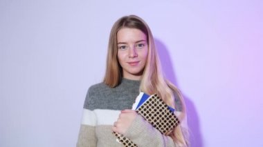 Güzel sarışın bir öğrencinin yakın plan fotoğrafı, elinde defterler, kameraya bakıyor ve arka plandaki parlak ışıkla gülümsüyor..