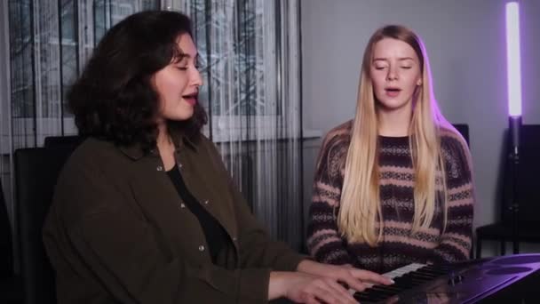 Dos chicas hermosas cantando una canción al piano. El profesor de música y el estudiante están analizando una nueva canción, dando una lección vocal. — Vídeos de Stock