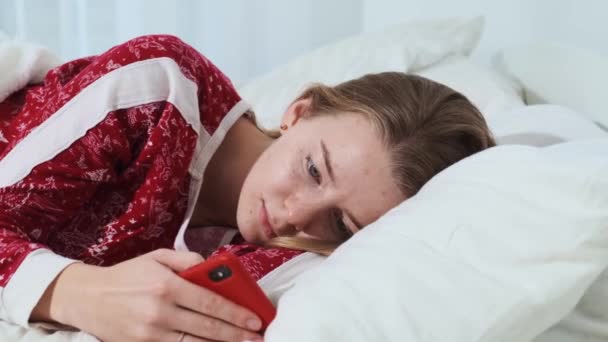 女の子は彼女の赤いパジャマで快適で居心地の良い、新鮮なベッドで彼女の手の中に彼女の携帯電話で、健康的で強い一日を楽しんでいます. — ストック動画