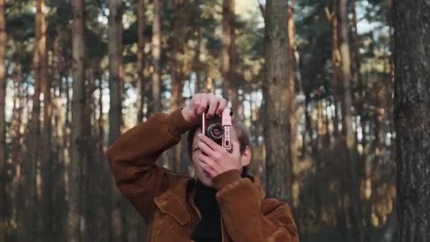 Młody blondyn fotograf robi zdjęcia natury. Fotograf robi zdjęcia w lesie aparatem retro. Młody fotograf ubrany w stylu retro. — Wideo stockowe