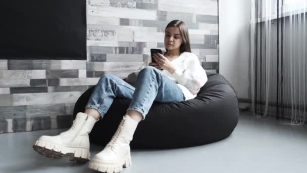 A girl is on her phone, sitting on a cushioned chair in a bright room. A girl writes a message using her phone. — Stock Video