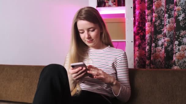 Retrato de uma menina que se senta no sofá e é interrompido por seu telefone. Uma menina digitando mensagens em seu telefone. — Vídeo de Stock