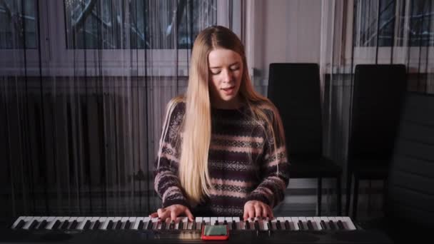 Uma jovem com longos cabelos brancos toca piano e canta uma canção. A menina está trabalhando em seus vocais e tocando piano. — Vídeo de Stock