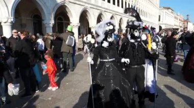 Siyah karnaval kıyafetleri ve maske takan bir çift, arka planda gezinen insanlar.