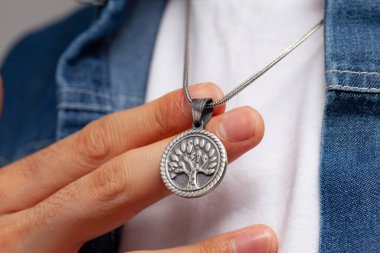 Kot ceketli ve beyaz tişörtlü karizmatik bir adam gri arka planda duruyor. Boynunda şık bir kolye var..