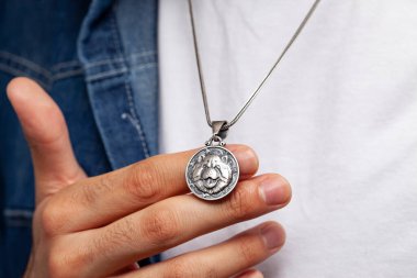 Kot ceketli ve beyaz tişörtlü karizmatik bir adam gri arka planda duruyor. Boynunda şık bir kolye var..