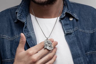 Kot ceketli ve beyaz tişörtlü karizmatik bir adam gri arka planda duruyor. Boynunda şık bir kolye var..